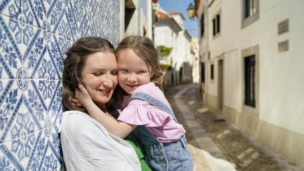 Mutter und Tochter umarmen sich lachend in einer sonnigen Gasse mit blauen Fliesen.