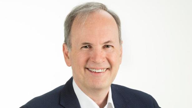 Mann mittleren Alters mit grauen Haaren trägt ein dunkelblaues Sakko und weißes Hemd und lächelt freundlich.