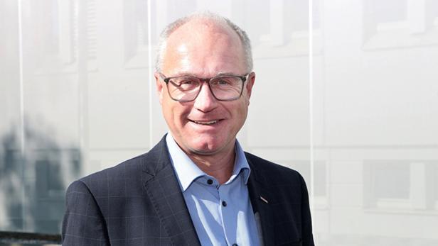 Ein älterer Mann mit Brille und Anzug lächelt vor einem hellen, unscharfen Hintergrund.