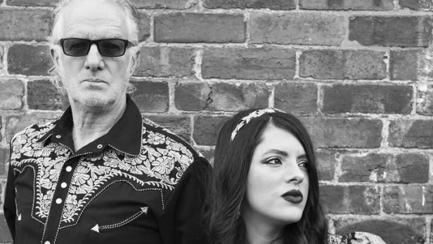 Ein älterer Mann mit Sonnenbrille und eine junge Frau mit langen Haaren stehen vor einer Backsteinmauer und schauen ernst.