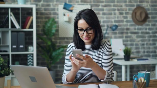 Frau mit Brille sitzt am Schreibtisch und nutzt ihr Smartphone in modernem Büro.