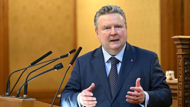 Ein Mann im Anzug spricht gestikulierend an einem Rednerpult mit mehreren Mikrofonen in einem holzverzierten Raum.