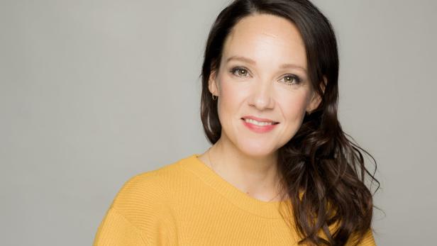 Eine lächelnde Carolin Kebekus mit braunen Haaren trägt einen gelben Pullover vor einem grauen Hintergrund.