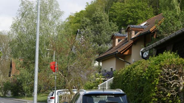 Ein Wohnhaus mit braunem Dach liegt an einer Straße mit parkenden Autos und viel Grün.