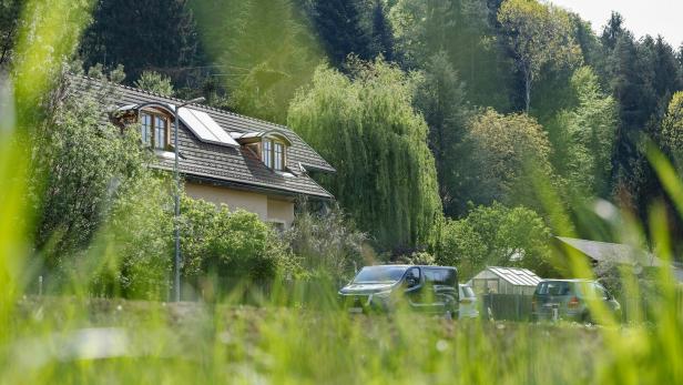 Ein Haus mit Dachfenstern steht vor einem bewaldeten Hügel, davor parken Autos.
