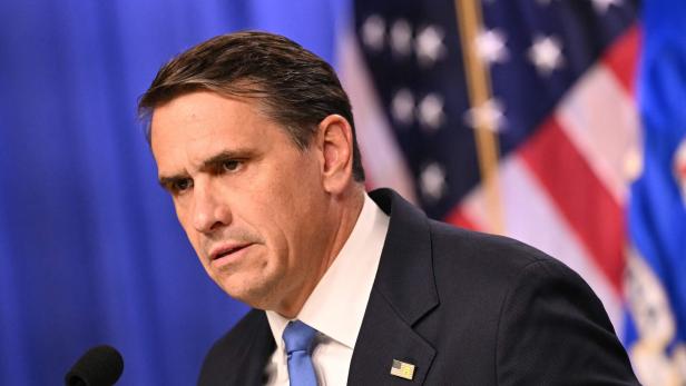 Ein Mann im Anzug mit blauem Hintergrund und einer US-Flagge spricht in ein Mikrofon.