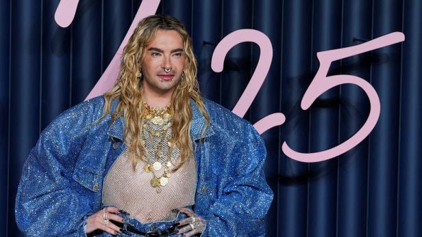 Bill Kaulitz mit blonden Locken, Nasenring und glitzernder Jeansjacke posiert vor blauem Hintergrund.