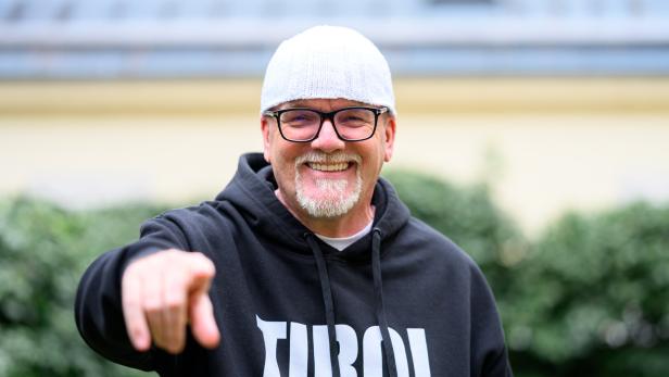 DJ Ötzi mit Brille und Mütze lächelt und zeigt auf den Betrachter, er trägt einen "Tirol"-Hoodie.
