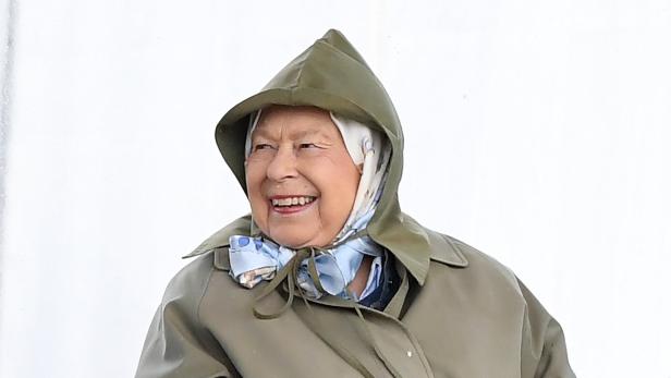 Königin Elizabeth II. lächelt in einem grünen Regenmantel mit Kapuze und einem hellblauen Halstuch.
