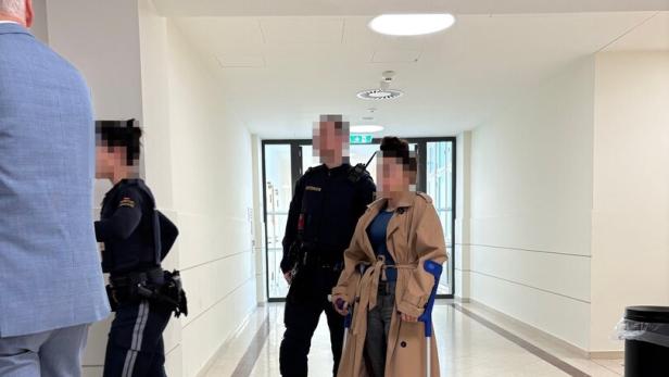 Eine Person mit Krücken und beigem Mantel steht neben Polizisten in einem hellen Flur.