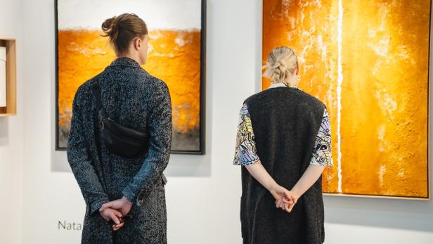 Zwei Personen betrachten abstrakte Gemälde in warmen Orangetönen an einer weißen Galeriewand.