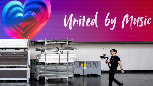 Eurovision Song Contest 2026 Ein Mann mit Kamera läuft vor einer großen bunten Herzgrafik und dem Schriftzug „United by Music“.