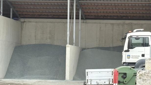 Zwei Haufen Streusalz oder Splitt unter einem Dach, daneben ein Kommunalfahrzeug mit Anhänger und Müllcontainer.