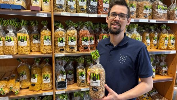 Ein Mann mit Brille steht vor Regalen voller verschiedener Nudelsorten und hält eine Packung Nudeln in der Hand.