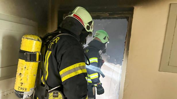Zwei Feuerwehrleute mit Atemschutzgeräten betreten einen verrauchten Raum und leuchten mit einer Taschenlampe hinein.