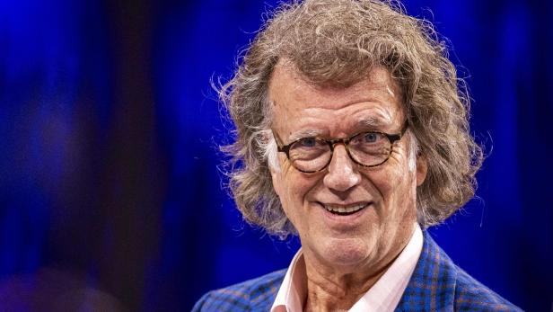 Ein lächelnder André Rieu mit Brille und kariertem Sakko vor blauem Hintergrund.