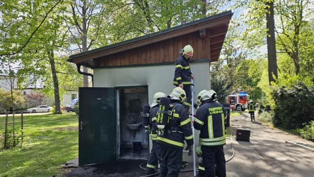 Feuerwehrleute untersuchen ein kleines Gebäude mit offener Tür und Brandschäden.