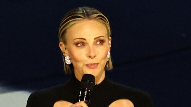 Charlize Theron mit blonden Haaren spricht in einem schwarzen Kleid mit Herz-Ausschnitt in ein Mikrofon.