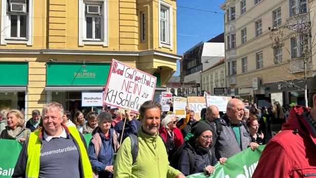Eine Gruppe von Menschen demonstriert mit Transparenten und Schildern auf einer sonnigen Straße vor gelben Gebäuden.