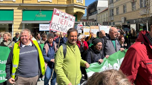 Eine Gruppe von Menschen demonstriert mit Transparenten und Schildern auf einer sonnigen Straße vor gelben Gebäuden.