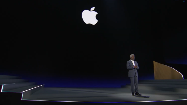 ein Mann im grauen Anzug auf einer Bühne, auf dem dunklen Hintergrund ist das Apple-Logo zu sehen