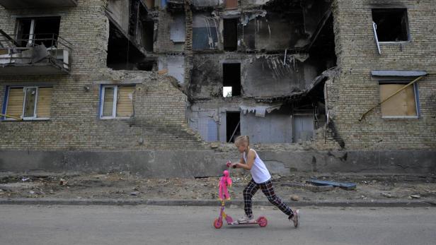 Ein Kind fährt auf einem Scooter, im Hintergrund ein zerbombtes Haus