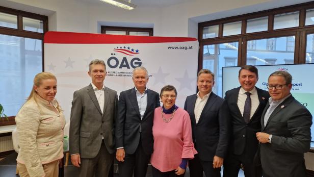 Sieben Personen stehen nebeneinander und lächeln in einem Raum mit ÖAG-Logo und großen Fenstern im Hintergrund.