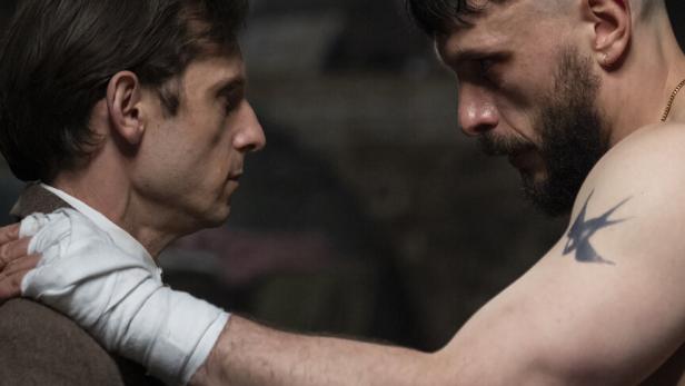 Niall und Ruben stehen sich angespannt gegenüber, einer trägt einen Anzug, der andere ist oberkörperfrei mit tätowiertem Arm.