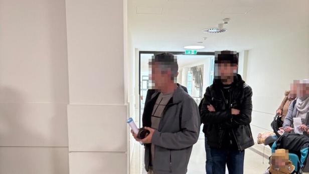 Mehrere Personen gehen durch einen hellen Flur, darunter ein Mann mit Dokumenten und ein Kind im Buggy.