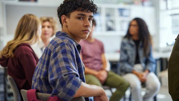 Ein Jugendlicher mit kariertem Hemd sitzt in einer Gruppe und blickt ernst zur Kamera, andere Jugendliche sind im Hintergrund.