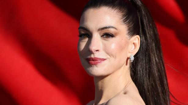 Anne Hathaway mit hochgesteckten, dunklen Haaren und Ohrringen vor rotem Hintergrund.