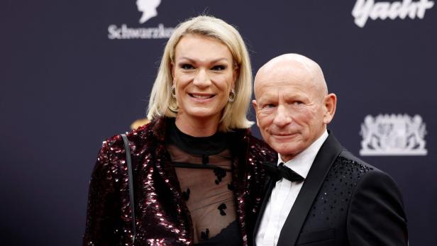 Maria Riesch mit blondem Haar und Johann Schrempf im Anzug posieren gemeinsam vor einem dunklen Hintergrund mit Logos.