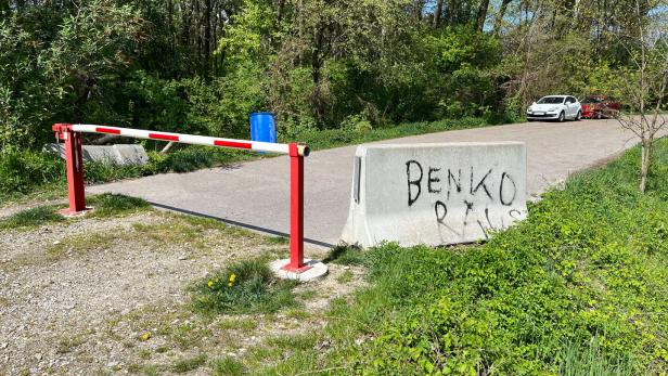 Eine rot-weiße Schranke und eine Betonbarriere mit Graffiti versperren eine Straße im Grünen, im Hintergrund stehen zwei Autos.