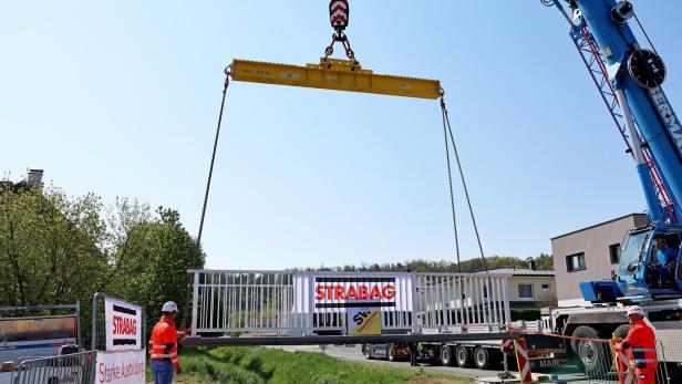 Ein Kran hebt eine weiße Brücke mit STRABAG-Logo, während zwei Bauarbeiter in Warnkleidung den Bereich sichern.