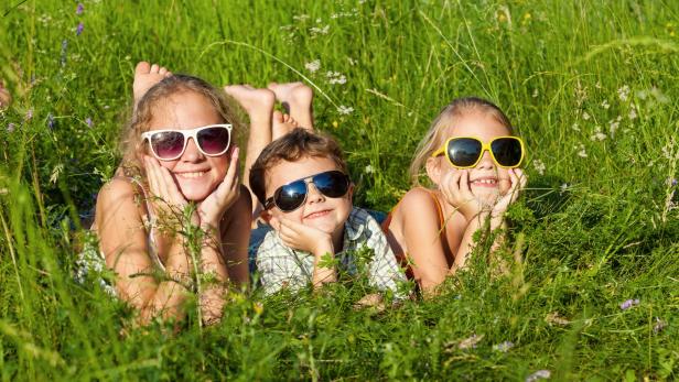 Das Bild zeigt drei fröhliche Kinder mit Sonnenbrillen in einer Wiese liegend.
