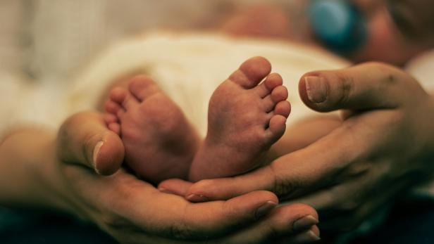 Erwachsene Hände halten sanft die Füße eines Babys, im Hintergrund ein Schnuller.