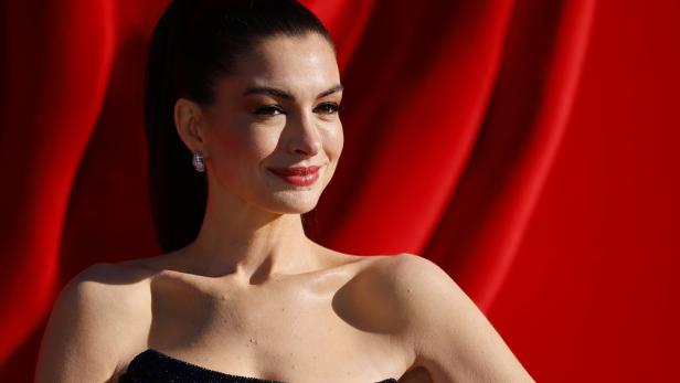 Anne Hathaway lächelt vor einem roten Hintergrund in einem trägerlosen, dunklen Kleid.