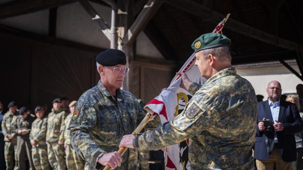 Zwei Soldaten in Uniform übergeben eine Fahne vor einer Gruppe von Personen.