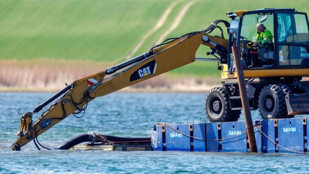 Ein Bagger mit langem Ausleger arbeitet auf einem Ponton im Wasser, ein Arbeiter sitzt in der Kabine.