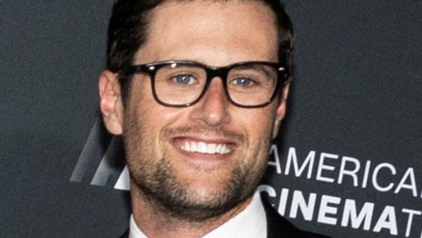 Ein Mann mit Brille, Anzug und Krawatte lächelt vor einer dunklen Wand mit weißer Schrift.