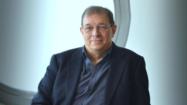 Ein Mann mit Brille und dunklem Sakko sitzt entspannt auf einem Stuhl vor einem hellen, modernen Hintergrund.