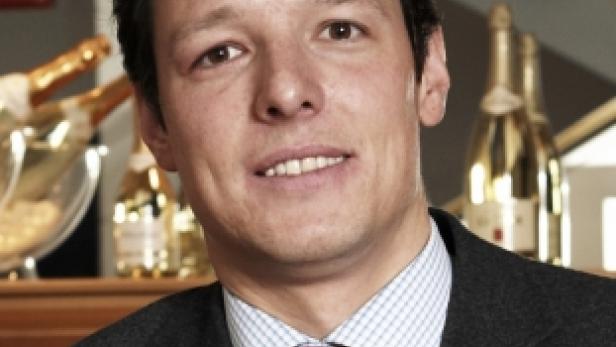 Ein Mann im Anzug mit Krawatte lächelt in die Kamera. Im Hintergrund sind Champagnerflaschen zu sehen.
