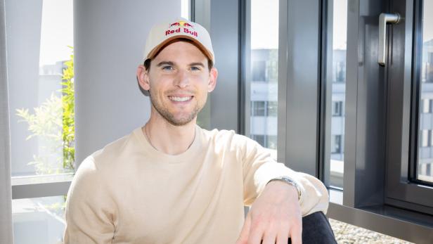 Ein junger Mann mit beigem Pullover und Red Bull-Kappe sitzt lächelnd vor einem Fenster mit modernem Rahmen.