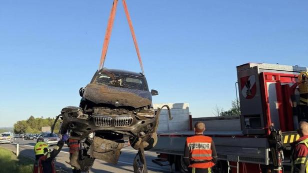 Ein stark beschädigtes Auto wird von einem Kran angehoben, während Feuerwehrleute daneben stehen.