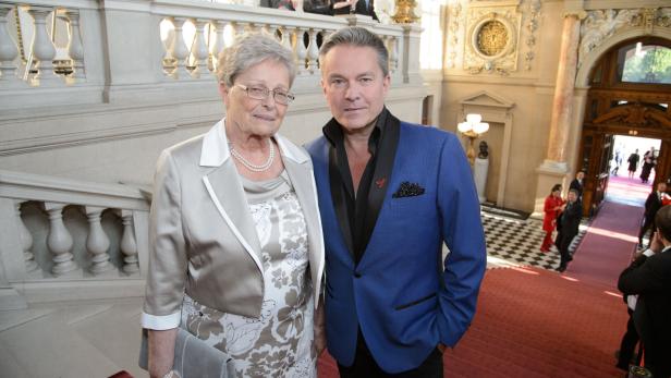 Eine ältere Frau und ein Mann im Anzug stehen auf einer prunkvollen Treppe in einem festlichen Gebäude.