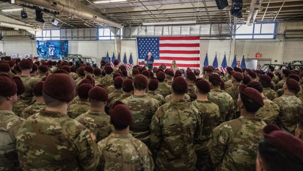 Eine Menschenmenge in Uniformen und roten Baretten hört einer Rede vor einer großen US-Flagge zu.