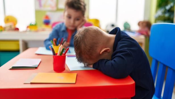Zwei Kinder sitzen an einem Tisch, eines legt den Kopf erschöpft auf die Arme, das andere malt mit Buntstiften.
