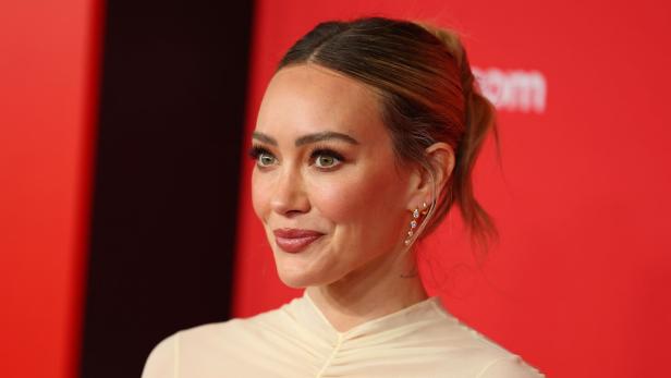 Hilary Duff mit hochgesteckten Haaren und Ohrringen vor rotem Hintergrund.