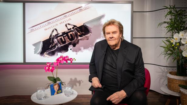 Howard Carpendale mit blondem Haar und schwarzem Sakko lächelt vor einem hellen Hintergrund.