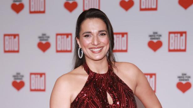 Eine lächelndeSally Özcan in einem roten, paillettenbesetzten Kleid posiert vor einem Hintergrund mit "Bild"-Logos und Herzen.